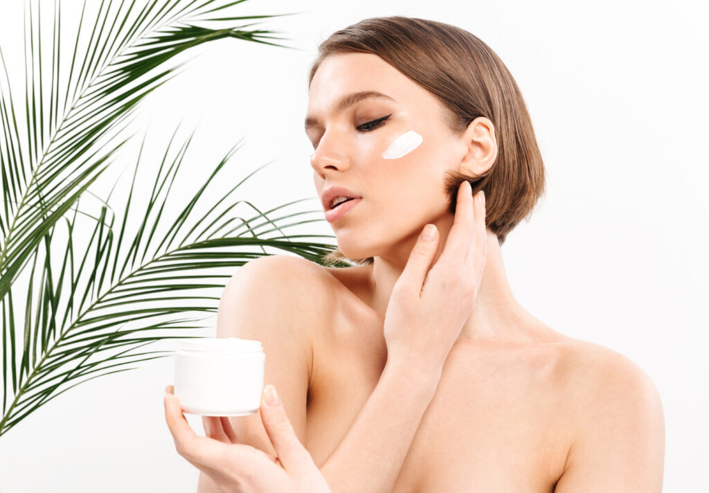 Beauty portrait of a lovely half naked woman with short brunette hair posing with green leaves and holding cream container isolated over white background